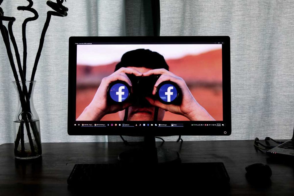 Tela de computador exibindo uma pessoa com um binóculo olhando o logo do facebook.